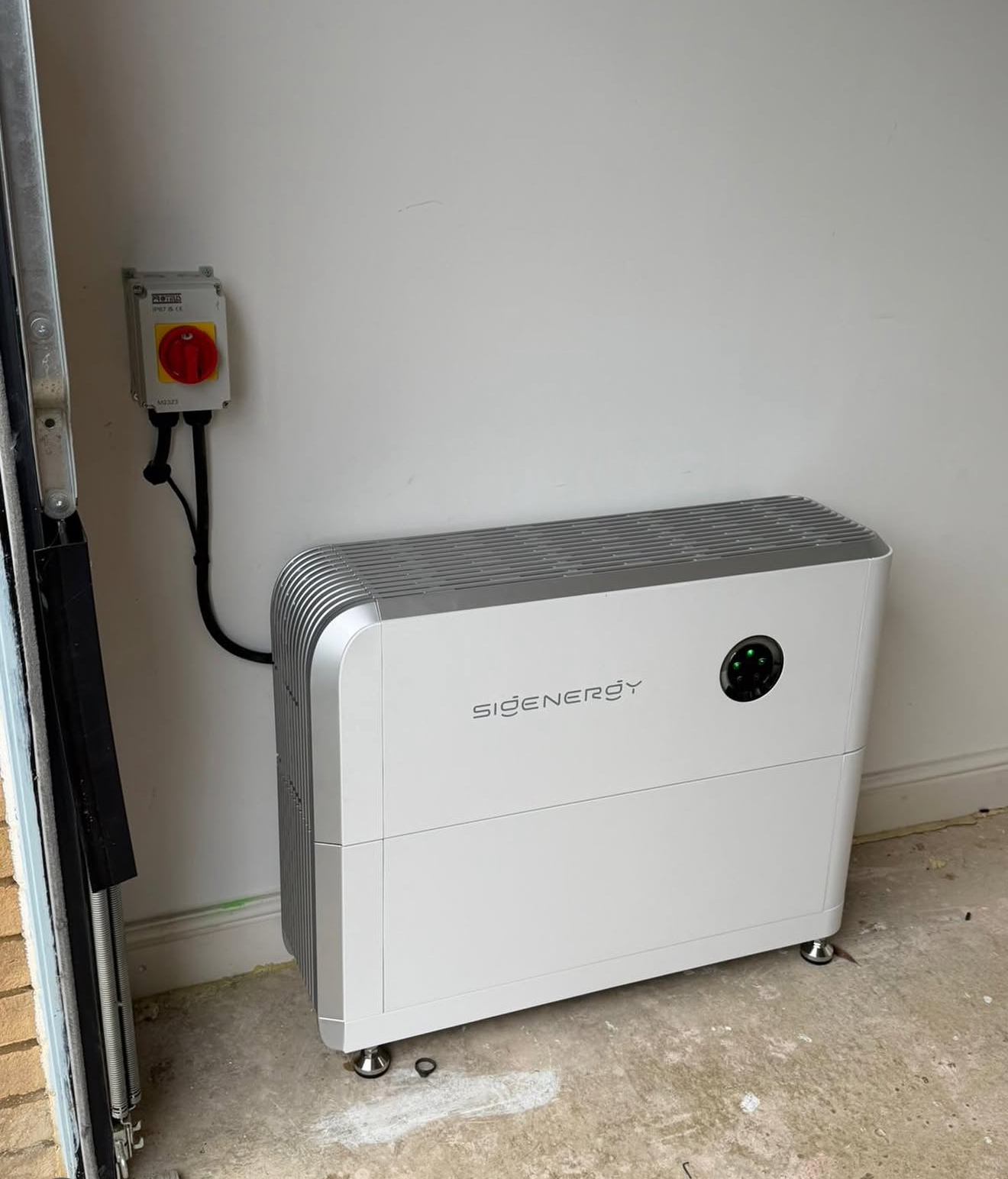 Floor-standing white Sigenergy home battery with a red safety isolator on the wall beside it, inside a garage